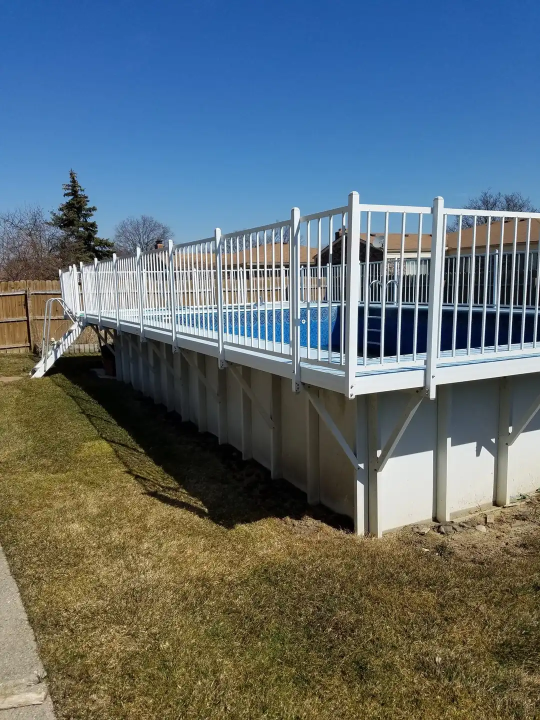 Long above-ground pool with a white aluminium railing deck and stairs, on a spring lawn under a clear sky