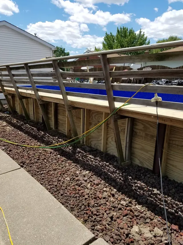Wood-braced pool wall with a deck railing above and a lava rock bed along the base